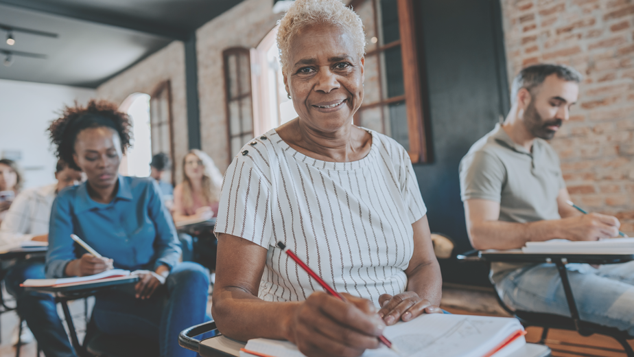 DC-ProgramHeaderBanner-iStock-1443628815 -924x520px A closeup of a matured aged smiling at the camera sitting in her diverse classroom of peers