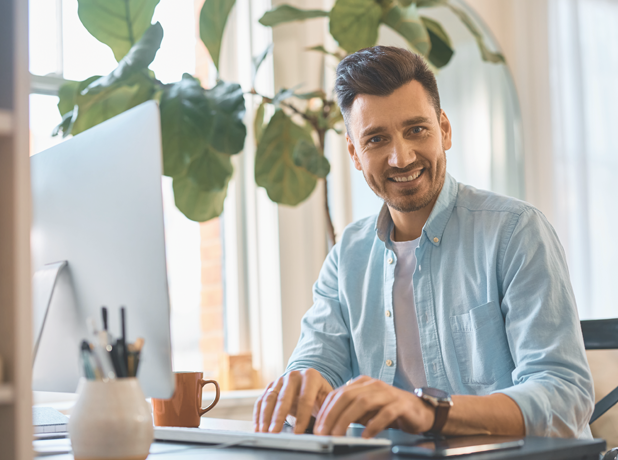 FinancialSupport-MediaTextSplitModule-618x460px-iStock-1457799390 Man at work in a casual office environment working on computer, looking at the camera and smiling.