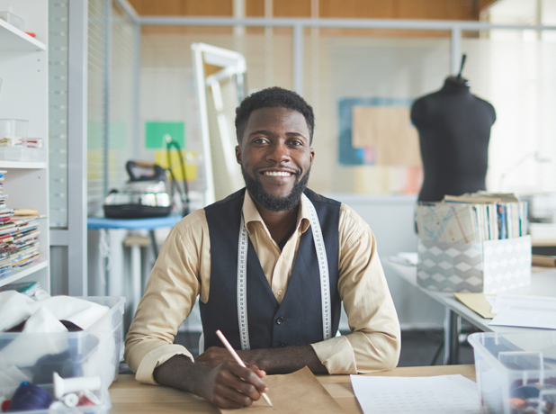 NDIS - Full Image - Media and Text Split 618x460px_iStock-1076098272 Young cheerful fashion designer sitting by table in his workshop and making new sketches in notepad.
