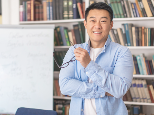 SEE-BodyImage-istock1470444547-618x460px Happy Asian Male Teacher Posing Holding Eyewear Standing In Classroom.