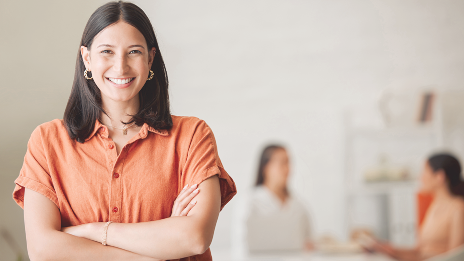 SmartandSkilled-HeaderBanner-924x520px-July24 Portrait of one confident young hispanic business woman standing with arms crossed in an office with her colleagues in the background. Ambitious entrepreneur and determined leader ready for success in a creative startup agency.