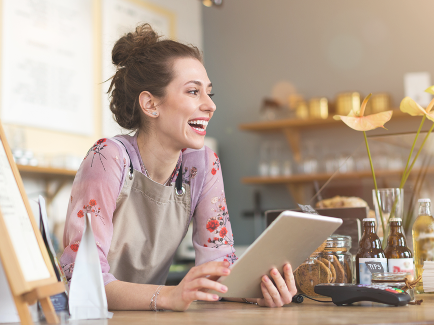 Skills-FullBleedBodyImage-iStock-1141489726 -858x643px Female shopkeeper smiling while using a digital tablet on shop counter.