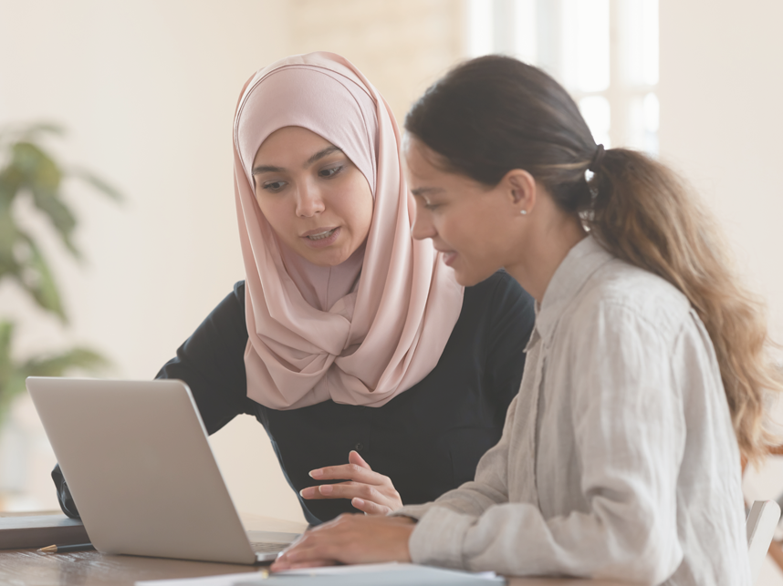 Support-FullImage - MediaandText SplitFull Bleed-858x643px-iStock-1192624591 Woman in hijab sitting with smiling colleague at table, looking at computer screen, explaining new support options.