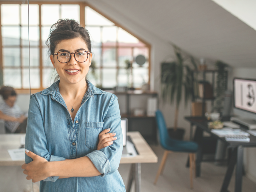 Support-FullImage - MediaandText SplitFull Bleed-858x643px-iStock-1350738281 Portrait of young smiling woman in creative office, standing with arms crossed, looking at camera.