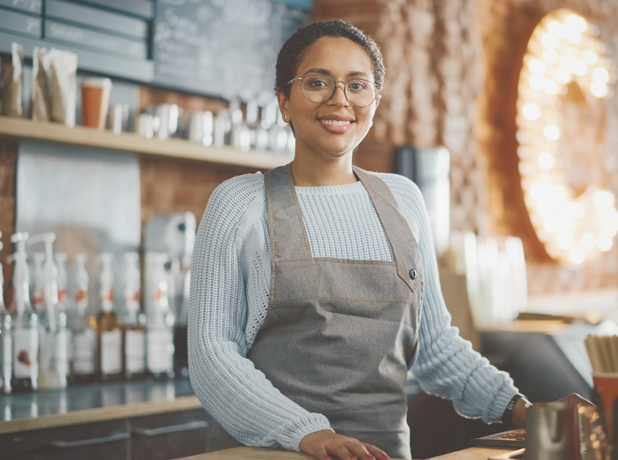 YEA - Full Image - Media and Text Split 618x460px_iStock-1293174927 Latin American female barista with short hair and glasses looking at camera and smiling in a coffee shop.