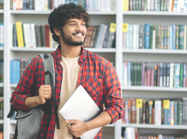 YEA - Full Image - Media and Text Split 618x460px_iStock-1419416580 Positive confident Middle-eastern male university student, in stylish casual wear, with backpack and laptop, stands in a library against the background of bookshelves.