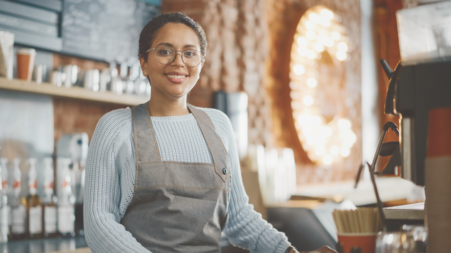 YEA-ProgramPageHeaderBanner-924x520-iStock-1293174927 Latin American female barista with short hair and glasses looking at camera and smiling in a coffee shop.