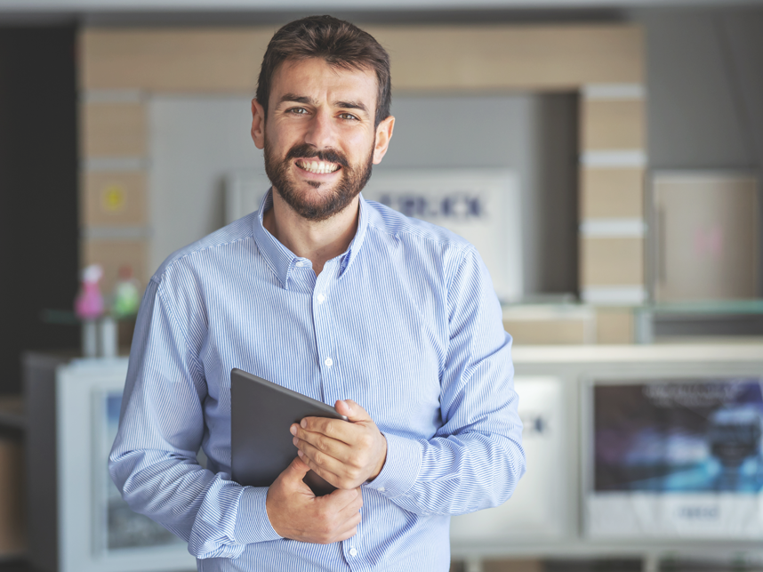 Employment-FullBleedModule-iStock-1284732216-858x643px Smiling bearded businessman standing in lobby of shipping firm and holding tablet while looking at camera.