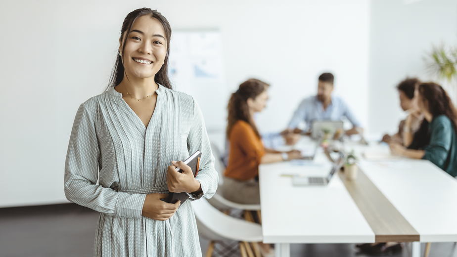 Financial Support-ProgramPageHeaderBanner-924x520px-iStock-1330547068 Confident young Asian businesswoman standing smiling at the camera in a boardroom with colleagues in the background.