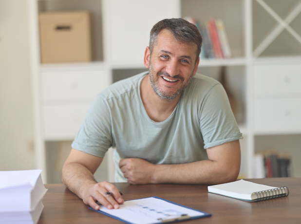 PBAS-BodyImageModule-iStock1677948942-618x460px Business man working at filling out documents at desk, looking at the camera and smiling.