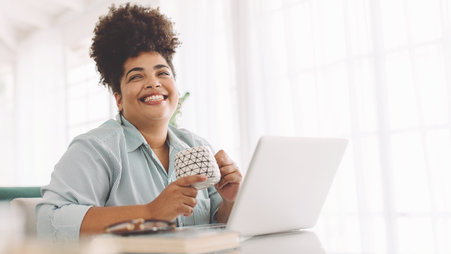 Program page -924x520px_1 Wide angle shot of happy African American woman sitting on table with coffee mug and laptop while working from home.
