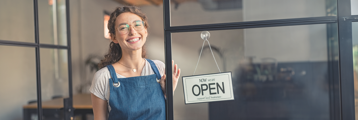 SEA-CampaignHeaderBanner-iStock-1289220719-1248x420px Youn,g joyful, curly-haired waitress with glasses standing at cafe entrance next to open for business sign.