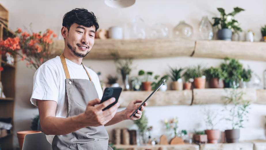 SEA- ProgramHeader Banner-iStock-1366720422-924x520px Asian male florist, owner of small business flower shop, using smartphone while working on digital tablet in work place. Checking stocks, taking customer orders, selling products online. Daily routine of running a small business with technology