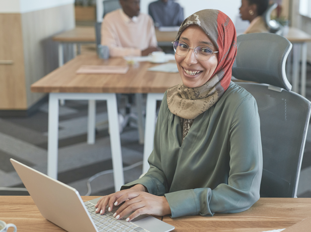 SEE -BodyImage-618x460px Smiling woman in headscarf and glasses sitting at desk on laptop