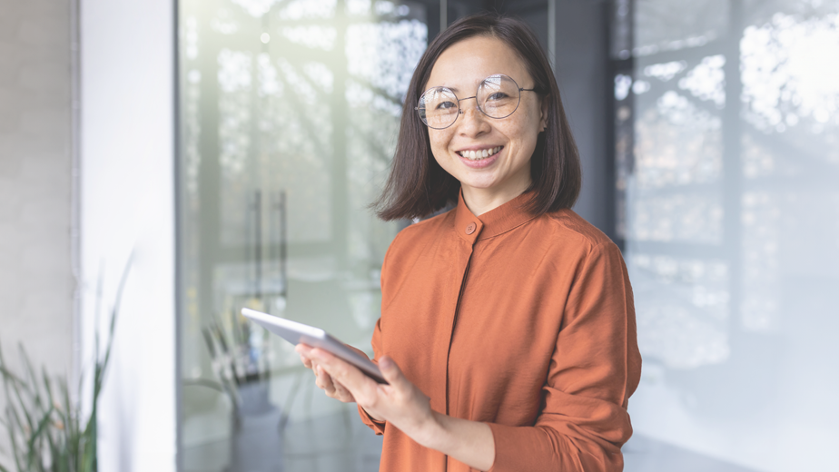 SEE- ProgramHeaderBanner-istock1492329923- 924x520px Portrait of a young Asian female with glasses smiling confidently to camera holding a touch screen device.