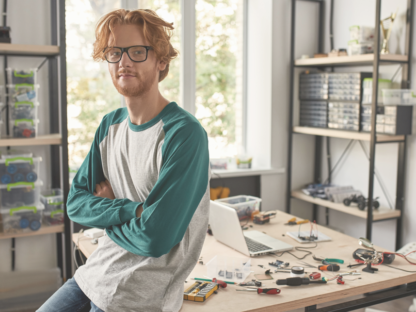 SQW-FullBleedModule-Istock1444716090-858x643px Male red haired Caucasian technician leaning on desk with crossed arms looking at camera in office. There are a lot of tools and equipment on the desk.