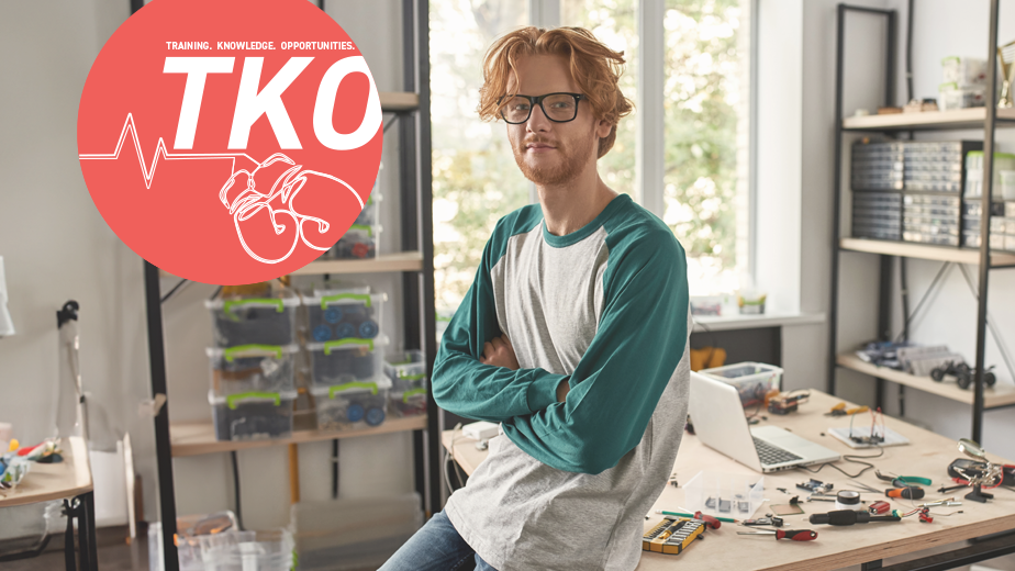 SQW-ProgramHeaderwithLogo-iStock1444716090-924x520px Male red haired caucasian technician leaning on desk with crossed arms looking at camera in office. There are a lot of tools and equipment on the desk.