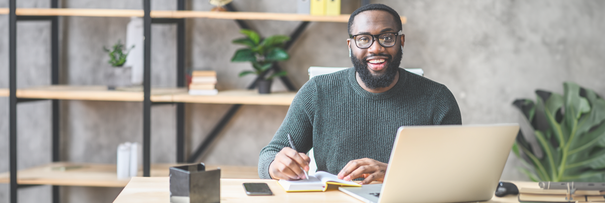 SS - Campaign Header Banner -1248x420px - Enrollment-updated Cheerful African-American male student or worker is watching online lectures or webinars and writing notes in a notebook.