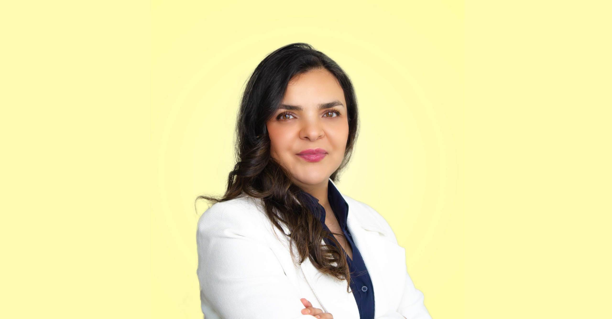 Samineh with arms folded in front of a pale yellow wall, wearing a white suit.